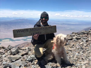 Smiling male in beanie hat and sunglasses crouching next to large blond fluffy dog on rocky mountain peak with a valley and distant mountains in the background.