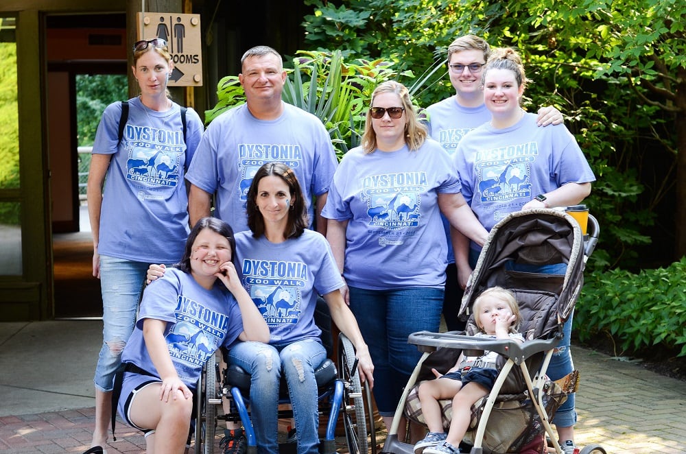 Five adults and two children, one in a stroller, posing for a photo. A young brunette woman in the front row is sitting in a wheelchair. All are wearing matching Dystonia Zoo Day t-shirts.