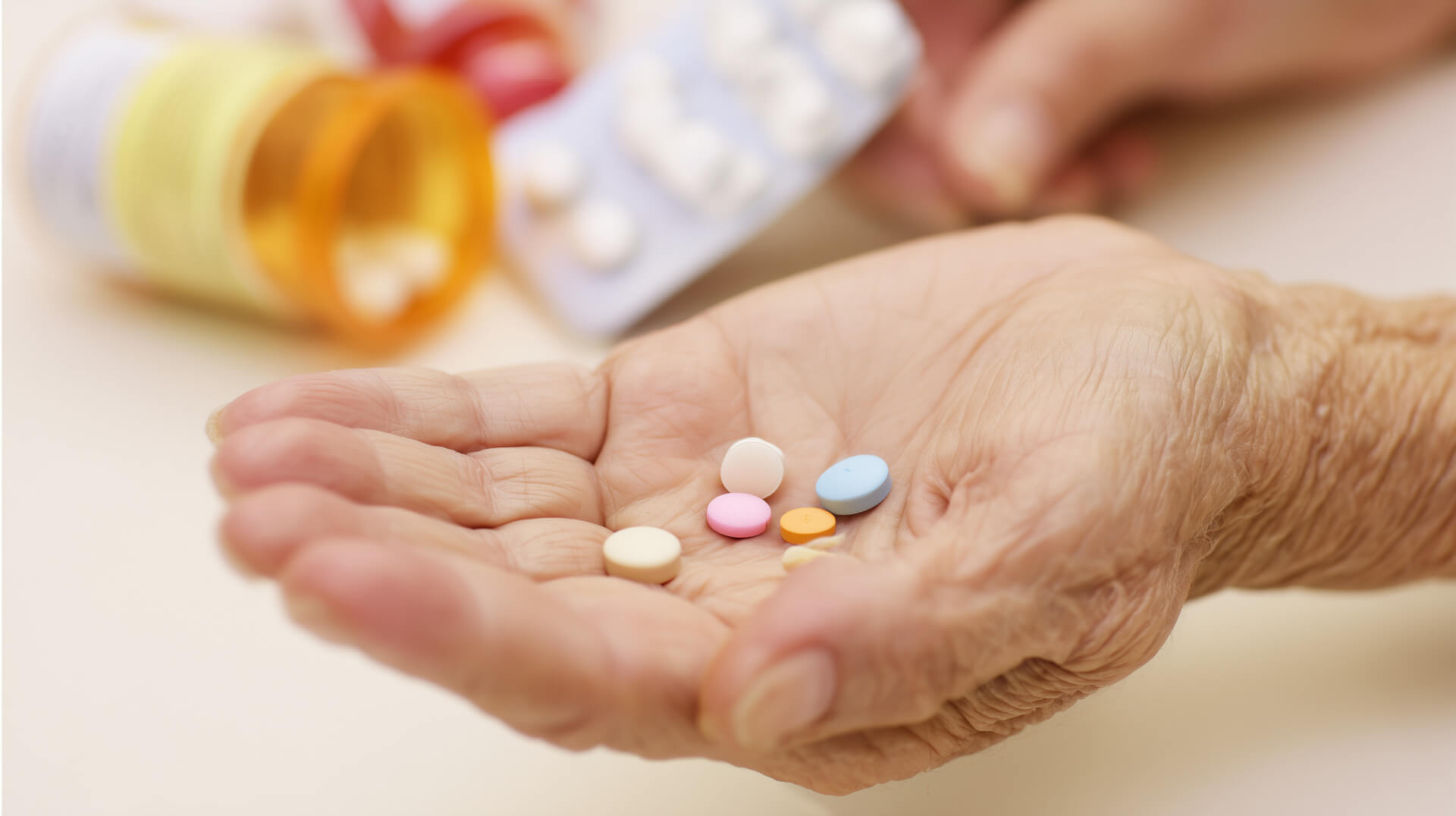 Close-up of an elderly hand holding multiple prescription pills with medicine bottle in the background.