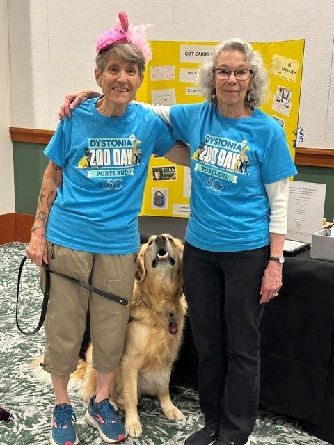 Two smiling seniors in matching blue 'Dystonia Zoo Day Portland' shirts pose with a golden retriever at an event, pink feather headband visible on the left person.