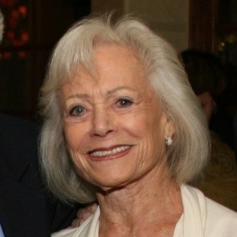 Smiling older woman with short gray hair and pearl earrings, wearing a light-colored blouse.