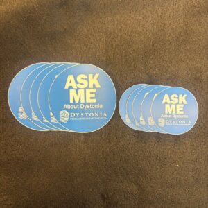 Blue circular stickers stacked on a dark carpet, reading 'ASK ME About Dystonia' with the Dystonia Medical Research Foundation logo.